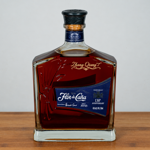A bottle of Fór de Cava Grand Cru 130th Anniversary rum, presented in a clear glass bottle with a dark amber liquid, a blue label, and a black cap, set on a wooden surface against a neutral gray background.