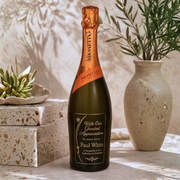 A customized bottle of Mionetto Prosecco stands on a stone surface in a styled setting. The dark green glass bottle features a gold foil top and a custom black label with gold text that reads 'With Our Greatest Appreciation For Your Support Paul White'.
