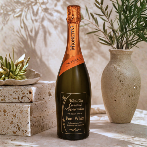 A customized bottle of Mionetto Prosecco stands on a stone surface in a styled setting. The dark green glass bottle features a gold foil top and a custom black label with gold text that reads 'With Our Greatest Appreciation For Your Support Paul White'.