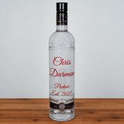 A clear glass bottle of vodka with a black cap and custom red text reading 'Chris Darmon Partner Est. 2027' on the front, standing on a wooden surface against a light gray background.