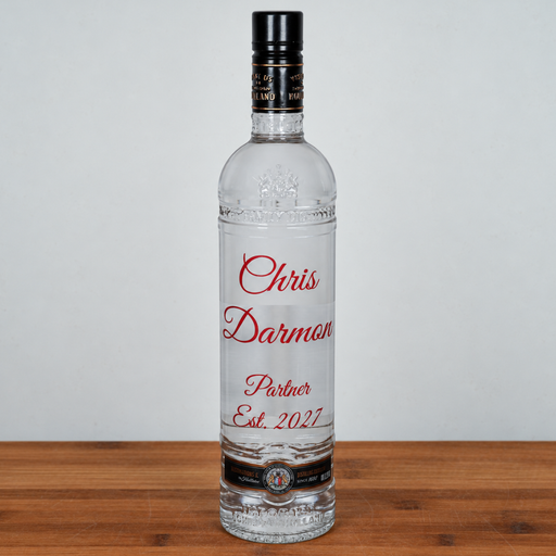 A clear glass bottle of vodka with a black cap and custom red text reading 'Chris Darmon Partner Est. 2027' on the front, standing on a wooden surface against a light gray background.