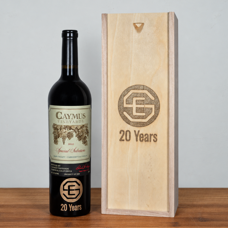 A bottle of Caymus Vineyards 20 Years red wine stands next to a light natural wood presentation box engraved with the same '20 Years' logo and monogram, set on a wooden surface against a light gray wall.