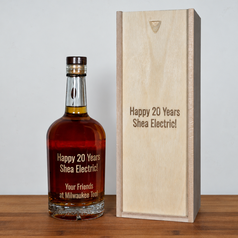 A bottle of amber liquor or whiskey stands beside a tall light wood box on a wooden surface against a plain light gray wall. Both pieces are engraved with the text 'Happy 20 Years Shea Electric!' and a smaller line below on the bottle, 'Your Friends at Milwaukee Too!'.