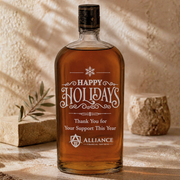 A clear glass bottle of amber liquor with a black cap and a large white holiday message on the front reading “Happy Holidays Thank You for Your Support This Year” and “Alliance Financial Partners,” photographed on a stone surface with warm shadows and small decorative props.
