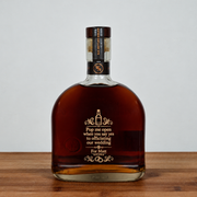A custom engraved bottle of whiskey with gold lettering on a dark brown glass body, set on a wooden surface against a light gray background.