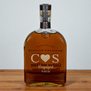 A bottle of Woodford Reserve bourbon whiskey with a custom-engraved 'C O S' heart design on the front, resting on a wooden surface against a neutral gray background.