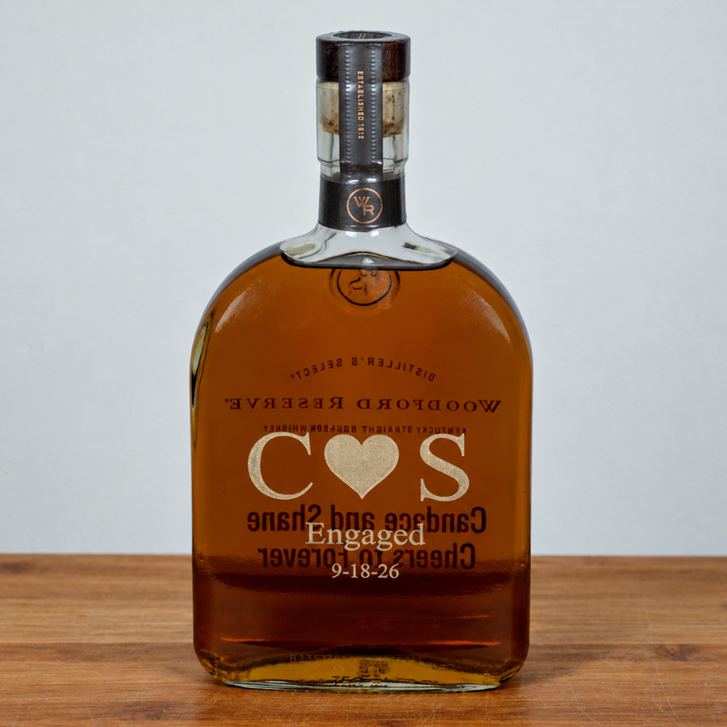 A bottle of Woodford Reserve bourbon whiskey with a custom-engraved 'C O S' heart design on the front, resting on a wooden surface against a neutral gray background.