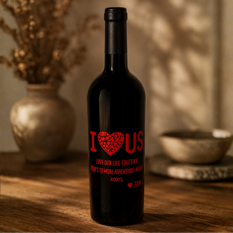 A dark glass bottle with a black cap stands on a wooden table in a warm, softly lit setting. The bottle features a red printed design that reads 'I [heart] US' with a patterned heart and smaller red text below, alongside a small photo placeholder area.