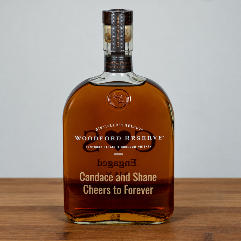 A custom-engraved bottle of Woodford Reserve Kentucky Straight Bourbon Whiskey on a wooden surface against a neutral gray background. The bottle features custom names, 'Candace and Shane', and the phrase 'Cheers to Forever' engraved on the front.