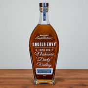 Angels Envy Triple Oak Neshanic Deutz Valley whiskey bottle on a wooden surface against a light gray background.