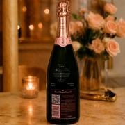 A bottle of Veuve Clicquot Ponsard champagne stands on a marble countertop in a warmly lit indoor setting, with a lit candle and a vase of pink roses blurred in the background.