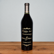 A dark glass caymus wine bottle with a metallic silver foil neck and a custom printed gold label celebrating a 2nd Anniversary, standing on a wooden surface against a light gray background.