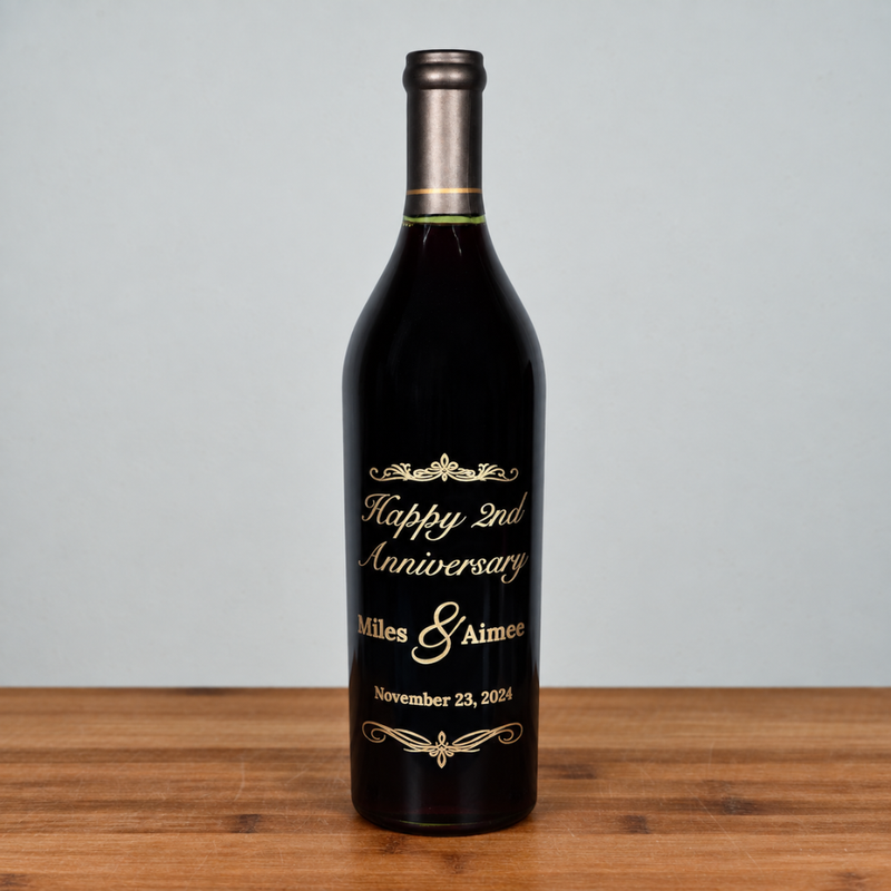 A dark glass caymus wine bottle with a metallic silver foil neck and a custom printed gold label celebrating a 2nd Anniversary, standing on a wooden surface against a light gray background.