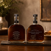 Two premium whiskey bottles displayed side by side on a dark wooden tray. The bottle on the left is labeled 'PHIL COLLINS SPECIAL RESERVE', and the bottle on the right is labeled 'WOODFORD RESERVE DOUBLE DALED'.