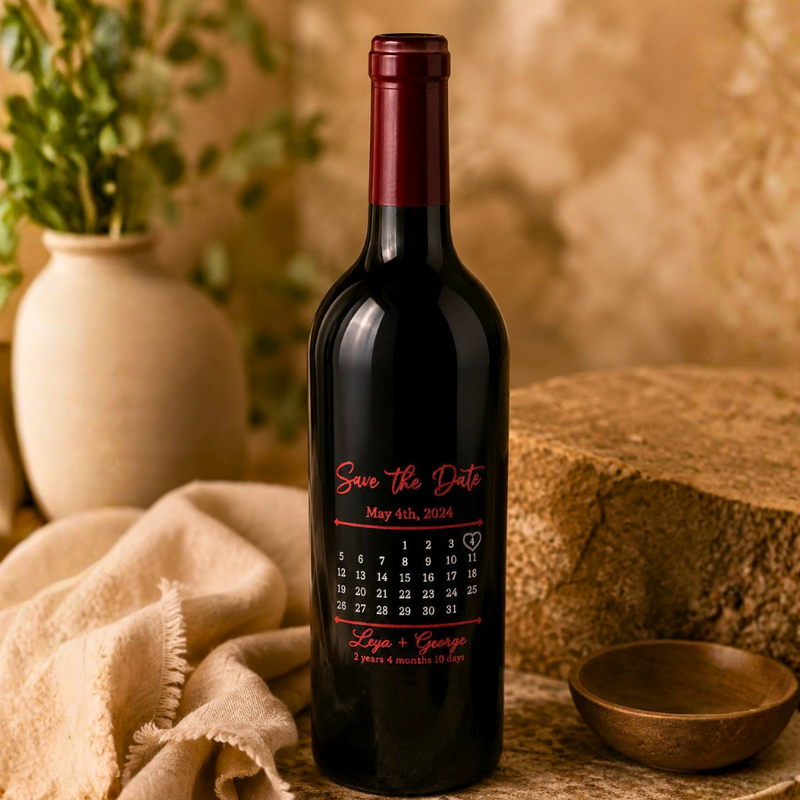 A dark wine bottle with a personalized 'Save the Date' label, styled on a rustic stone surface with a beige cloth, a small wooden bowl, and a vase of greenery in the background.
