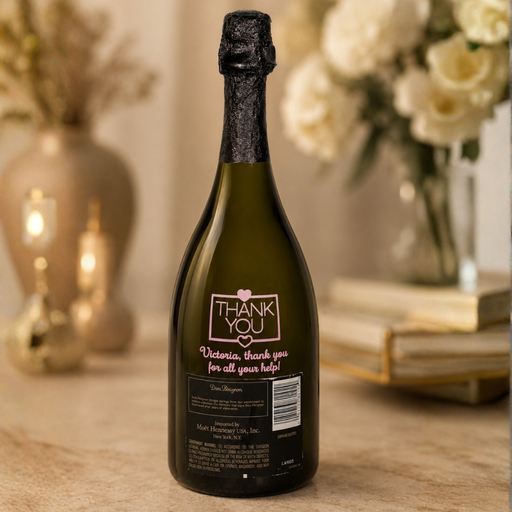 A dark green bottle of sparkling wine or champagne with a black foil top and a custom 'Thank You' label, set on a beige stone surface with a blurred background of flowers and candles.
