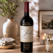 A bottle of Josh Red Label Cabernet Sauvignon red wine stands on a wooden table in a warmly lit, cozy room setting with a vase, framed picture, and candles in the background.