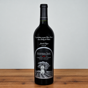 A single bottle of Silver Oak Cabernet Sauvignon red wine stands on a wooden surface against a light gray background. The bottle is dark glass with a black cap and a custom black label featuring silver and white text and an illustration of a vineyard.