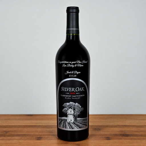 A single bottle of Silver Oak Cabernet Sauvignon red wine stands on a wooden surface against a light gray background. The bottle is dark glass with a black cap and a custom black label featuring silver and white text and an illustration of a vineyard.