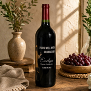 A dark glass wine bottle with a personalized graduation label stands on a wooden table in a warm, styled interior. The label reads 'PAIRS WELL WITH GRADUATION, Evelyn, Boston University, CLASS OF 2027' with a small illustrated figure.