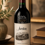 A custom wine bottle with a personalized label for 'Jordan', featuring a vintage-style illustration of a vineyard and the text 'And They Lived Happily Ever After' and 'Alan & Rebecca September 7, 2027'. The bottle is styled on a wooden surface with a blurred background containing books and a vase.