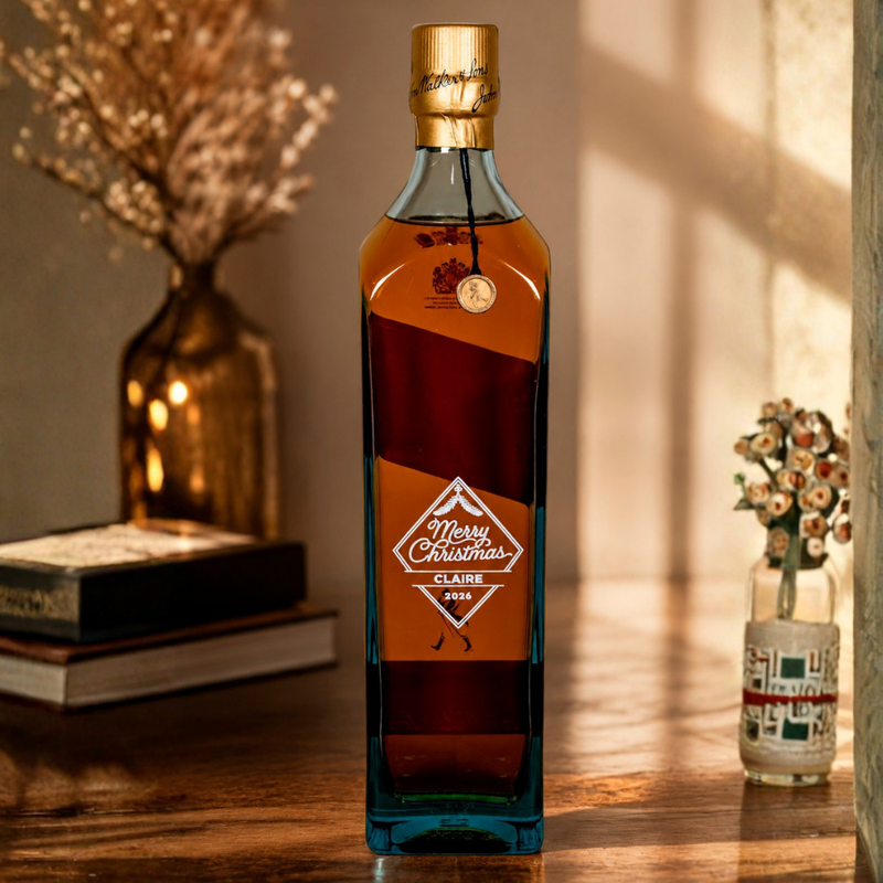 A square glass bottle of liquor with amber liquid and a gold cap, standing on a wooden table. The front label reads 'Merry Christmas 2024' with a red and orange design.