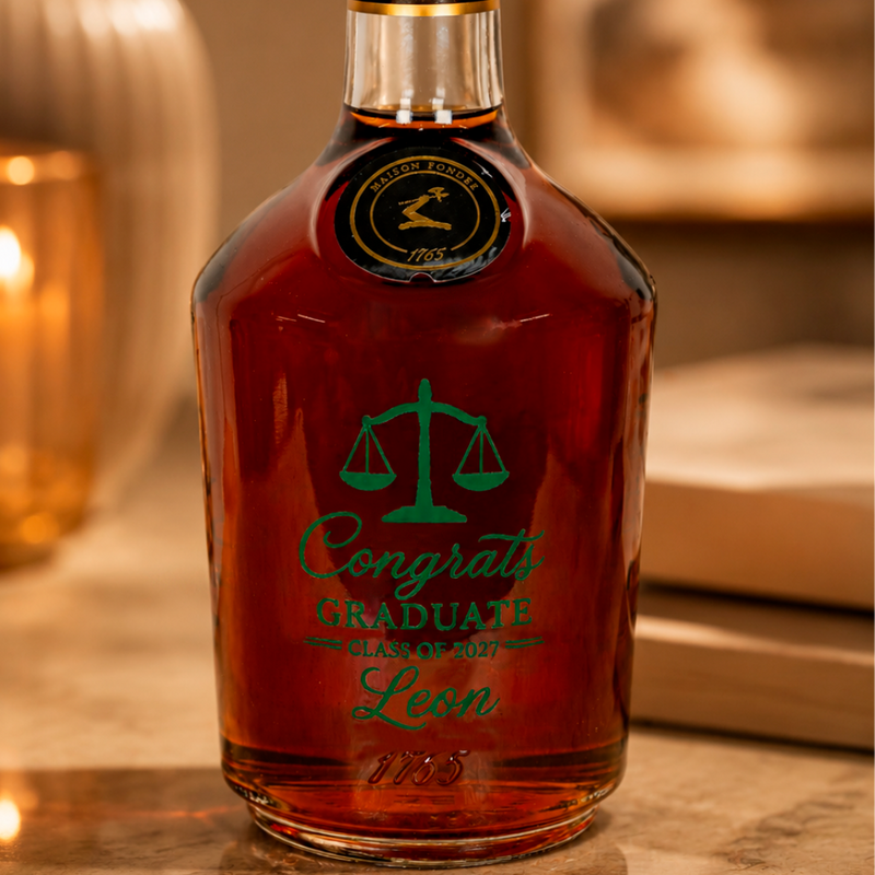 A bottle of amber liquor with a green engraved graduation design reading 'Congrats Graduate Class of 2027 Leon' and a gold seal near the neck, photographed on a warm blurred tabletop with candles in the background.