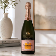 A bottle of Greystone Capital Veuve Clicquot Rosé champagne stands on a light marble surface in a softly lit interior setting, with a white vase and books blurred in the background.