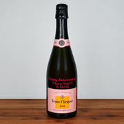 A bottle of Veuve Clicquot Rosé champagne with a custom pink anniversary label reading 'Happy Anniversary I Love You!!! 7/19/26' on a wooden surface against a plain light gray background.