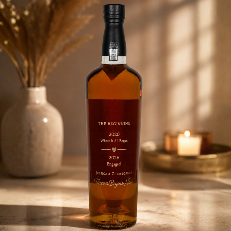 A personalized amber glass bottle with a black cap stands on a marble surface in a warmly lit room. The bottle is engraved with wedding-themed text including 'THE BEGINNING 2020 Where It All Began 2026 Engaged Jessica & Christopher Forever Begins Now.