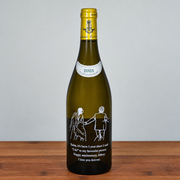 A green glass wine bottle with a gold foil top and a white printed illustration of a couple holding hands, with a humorous anniversary message below. The bottle is standing on a wooden surface against a plain light gray background.