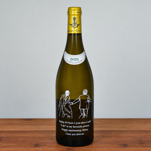 A green glass wine bottle with a gold foil top and a white printed illustration of a couple holding hands, with a humorous anniversary message below. The bottle is standing on a wooden surface against a plain light gray background.