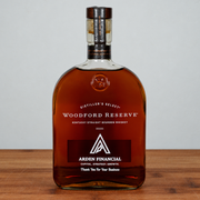 A bottle of Woodford Reserve Kentucky Straight Bourbon Whiskey with a custom Arden Financial graphic on the label, displayed on a wooden surface against a neutral gray background.