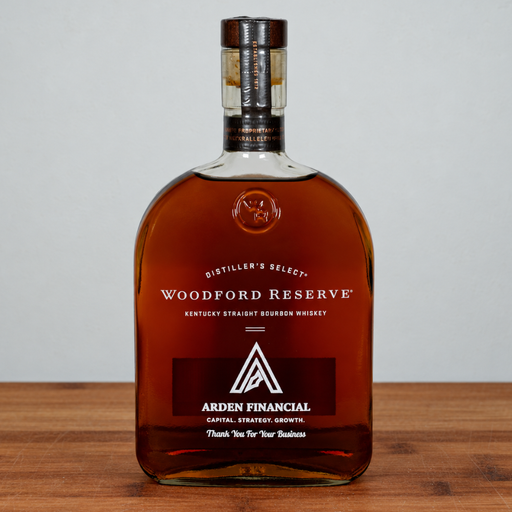 A bottle of Woodford Reserve Kentucky Straight Bourbon Whiskey with a custom Arden Financial graphic on the label, displayed on a wooden surface against a neutral gray background.