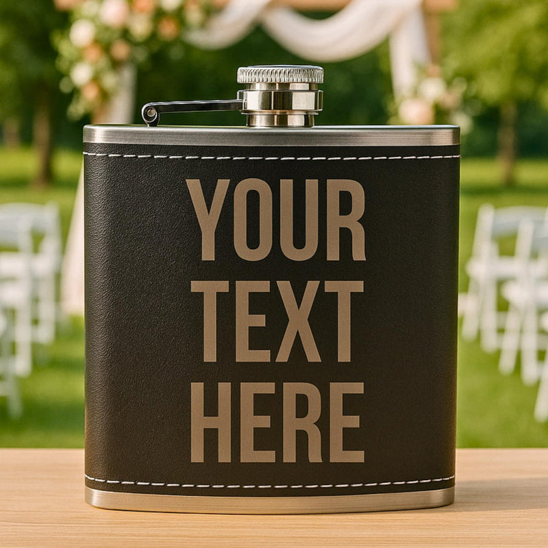 Personalized black leather flask with 'YOUR TEXT HERE' on a blurred outdoor background