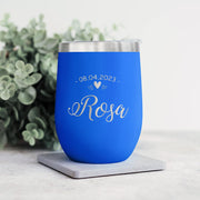 Blue wine tumbler with engraved text on a white surface with greenery in the background