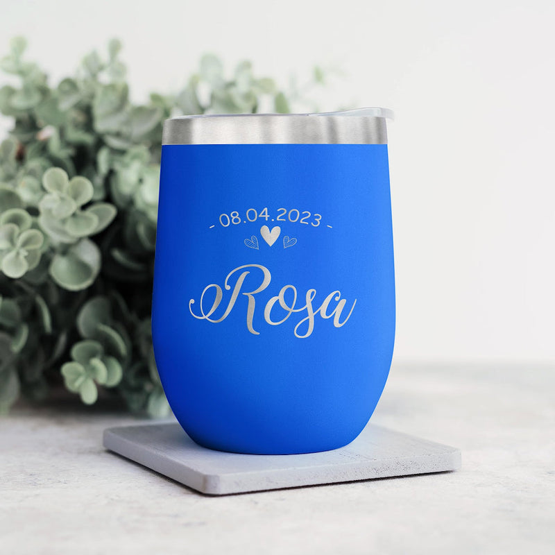 Blue wine tumbler with engraved text on a white surface with greenery in the background