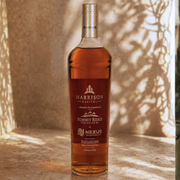 A bottle of Harrison Capital whiskey with a dark amber label and clear glass, standing on a stone surface with warm sunlight and leaf shadows cast across the background.
