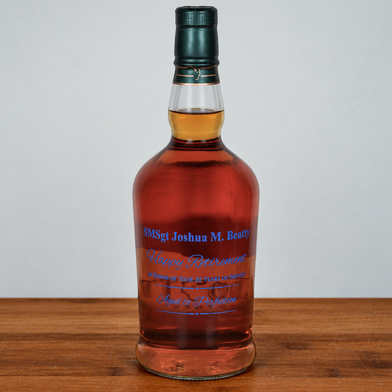 A bottle of amber liquor with blue script text on the front, sealed with a dark green cap, standing on a wooden surface against a light gray background.