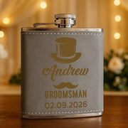 Gray flask with gold engraving of a top hat, mustache, and text on a wooden surface with blurred lights and flowers in the background.