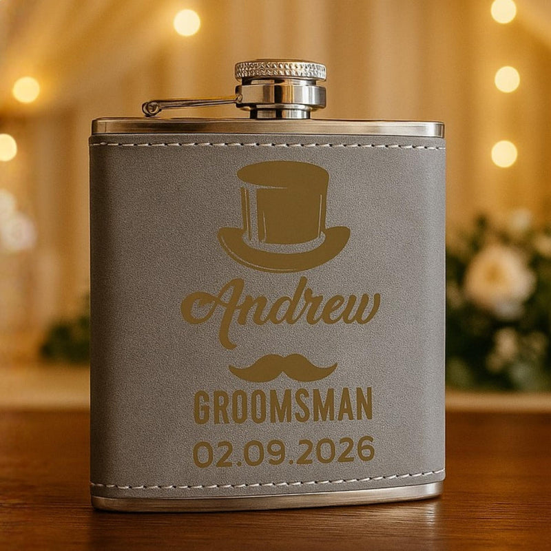 Gray flask with gold engraving of a top hat, mustache, and text on a wooden surface with blurred lights and flowers in the background.