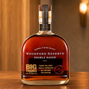 A bottle of Woodford Reserve Double Oaked bourbon whiskey on a wooden surface with a warm, blurred background. The bottle is filled with amber liquid and features a black label with gold and white text.