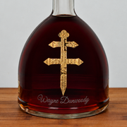 A clear congnac bottle filled with dark red liquid, featuring a large gold Orthodox cross emblem on the front and the name 'Wayne Dunwoody' in white script near the base, set on a wooden surface against a light gray background.