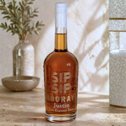 A personalized bottle of amber liquor with a white textured neck wrap, standing on a stone surface. The bottle features custom etched text reading 'SIP SIP MOORAY! Justin You Earned This'.