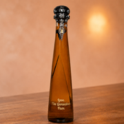An amber glass bottle with a black ornate cap and gold lettering that reads 'Love, The Gonsalves Fam' on a wooden surface against a warm beige background.