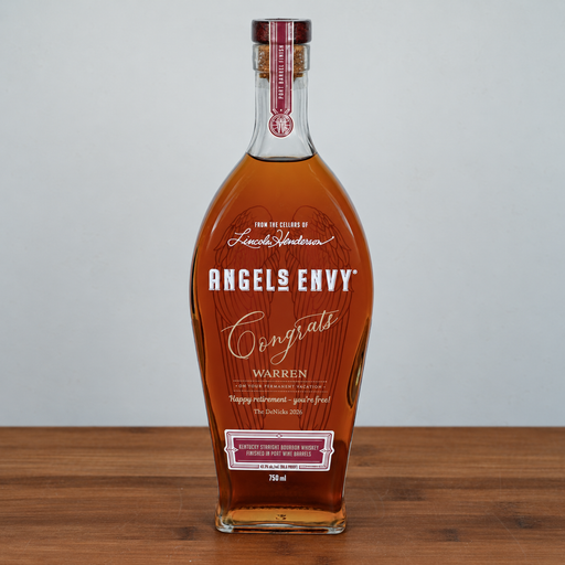 Angels Envy Congrats Warren cognac liqueur in a clear glass bottle with amber liquid, set on a wooden surface against a light gray background.