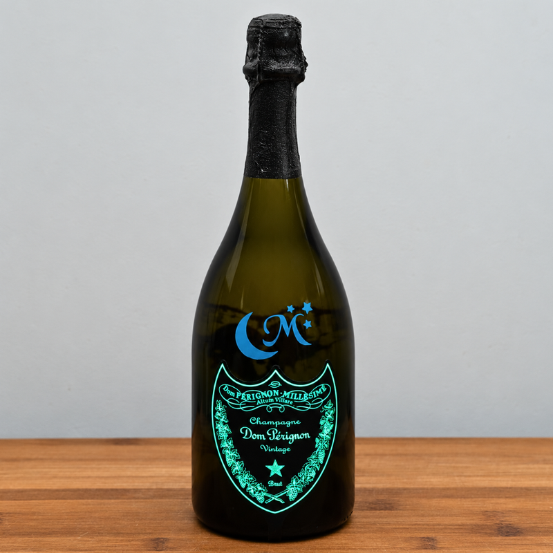 A dom perignon luminous champagne bottle with a black foil top and a glowing turquoise label, standing on a wooden surface against a light gray background.