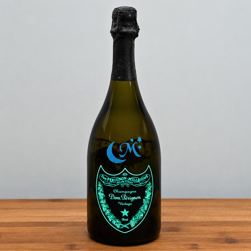 A dom perignon luminous champagne bottle with a black foil top and a glowing turquoise label, standing on a wooden surface against a light gray background.