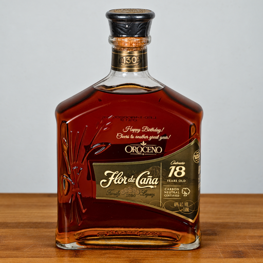 A bottle of Oroceno Flor de Cañ 18th birthday cognac, featuring a clear glass bottle filled with amber liquid, a black and gold cap, and a gold and black label, set on a wooden surface against a light gray background.