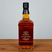 A customized Jack Daniel's 16 oz square glass whiskey bottle with amber liquid, featuring a black cap and a personalized 'Happy Anniversary Jacob & Lindsey' label, set on a wooden surface against a neutral gray background.